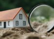 A miniature house sits on soil, examined through a magnifying gl